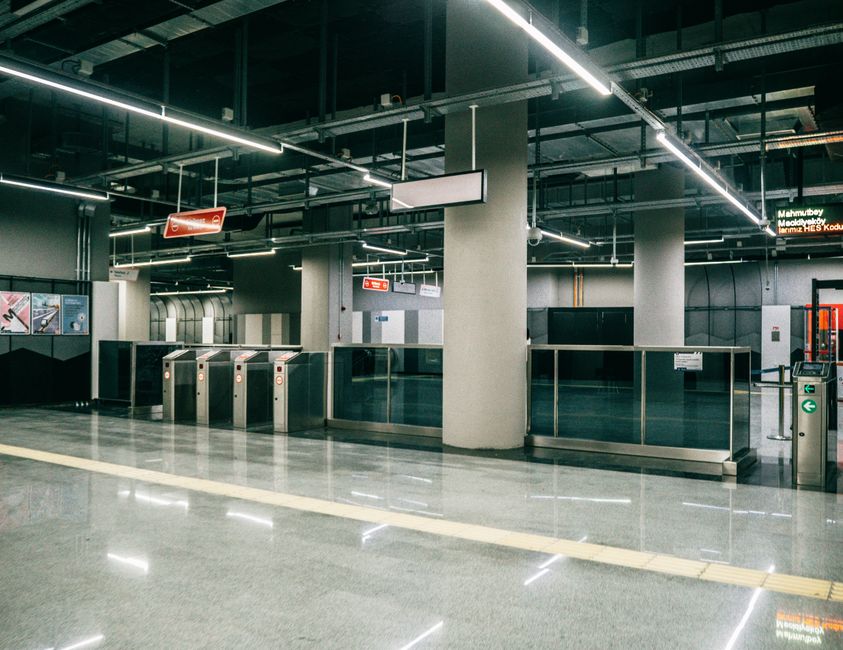 An empty, modern underground station entrance near Bow Road Station, featuring a polished gray stone floor with a highly reflective surface, illuminated by bright linear LED ceiling lights. The area includes stainless steel turnstiles with red and white card readers, glass barriers with metal frames, and a digital display board showing station information. Structural pillars in neutral tones support the ceiling, while clean glass panels enhance the sleek appearance. The space appears freshly cleaned, showcasing no dust or debris, with a clear, hygienic environment, reflecting the professional cleaning services offered by Cleaners E3 for public transportation areas.