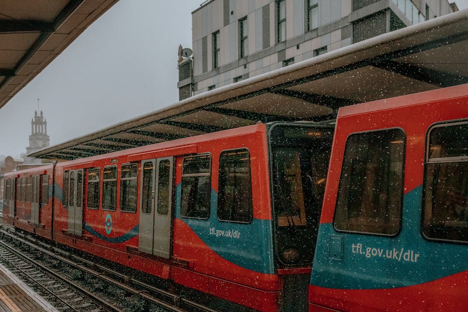 An exterior view of Bow Road Station showing a red and blue London Underground train parked on the tracks under a modern station canopy with a metal roof. Snowfall is visible, with snowflakes falling onto the train and platform. The station's platform is partially visible, with a building in the background featuring a tall clock tower. The lighting is natural and subdued, capturing a cold, winter day. [COMPANY_NAME] provides professional cleaning services including surface cleaning and sanitisation, ensuring hygiene and cleanliness for transportation hubs.