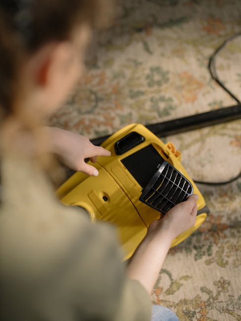 A person operating a yellow portable fan heater or dehumidifier on a patterned carpet in a room, likely during a surface cleaning or drying process. The device has a black vent grille on the front, and the person’s hand is pressing a button or adjusting the settings. The room is illuminated with natural or artificial light, highlighting the clean, plush carpet with floral or ornate designs. The scene demonstrates a focus on maintaining or restoring cleanliness and hygiene in a domestic or commercial setting, consistent with best practices for deep cleaning near Bow, SE3, as promoted by Cleaners E3.
