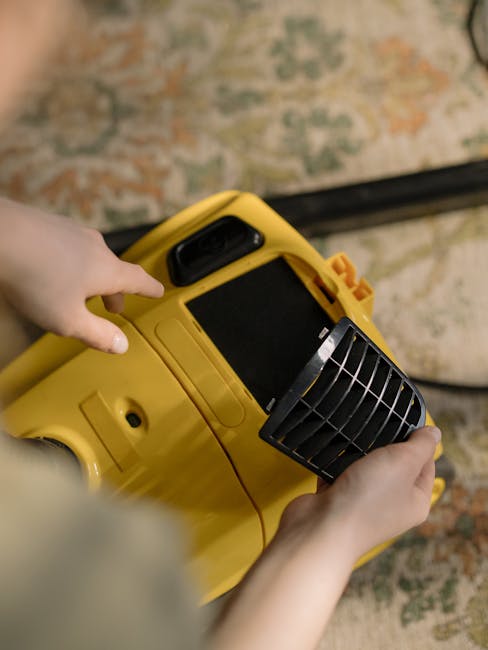 A person operating a yellow portable fan heater or dehumidifier on a patterned carpet in a room, likely during a surface cleaning or drying process. The device has a black vent grille on the front, and the person’s hand is pressing a button or adjusting the settings. The room is illuminated with natural or artificial light, highlighting the clean, plush carpet with floral or ornate designs. The scene demonstrates a focus on maintaining or restoring cleanliness and hygiene in a domestic or commercial setting, consistent with best practices for deep cleaning near Bow, SE3, as promoted by Cleaners E3.