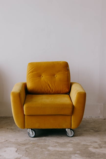 A mustard yellow upholstered armchair with padded armrests and button-tufted backrest sits on small caster wheels, positioned against a plain white wall in a room with a concrete floor. The surface of the chair appears clean and well-maintained, with no visible dust or stains. Natural light illuminates the scene, reflecting subtly on the fabric, indicating a tidy and fresh environment. This image showcases the importance of surface cleaning and maintenance in domestic spaces, aligning with Mile End Park upholstery cleaning tips for Bow residents, SE3, provided by Cleaners E3, specialists in professional cleaning services.