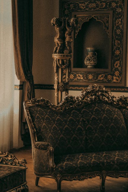 A richly decorated antique-style living room featuring ornate wooden furniture with intricate carvings and upholstered in dark, patterned fabric. The room includes a vintage sofa with curved armrests and a matching side table. Behind the sofa is an elaborate wall mirror with a gilded frame, displaying classical floral motifs, and a decorative ceramic vase sits on a shelf within the mirror's frame. To the left, heavy velvet curtains in a deep hue frame a window, allowing soft natural light to illuminate the space. The overall scene conveys a sense of traditional elegance, highlighting the importance of surface cleaning and maintenance in preserving such detailed furnishings. For residents seeking to maintain this level of cleanliness and appearance, Cleaners E3 offers specialised domestic cleaning services, including upholstery deep cleaning and upholstery sanitisation, tailored to protect and enhance vintage and luxurious furniture, as discussed in the Mile End Park upholstery cleaning tips for Bow residents, SE3.