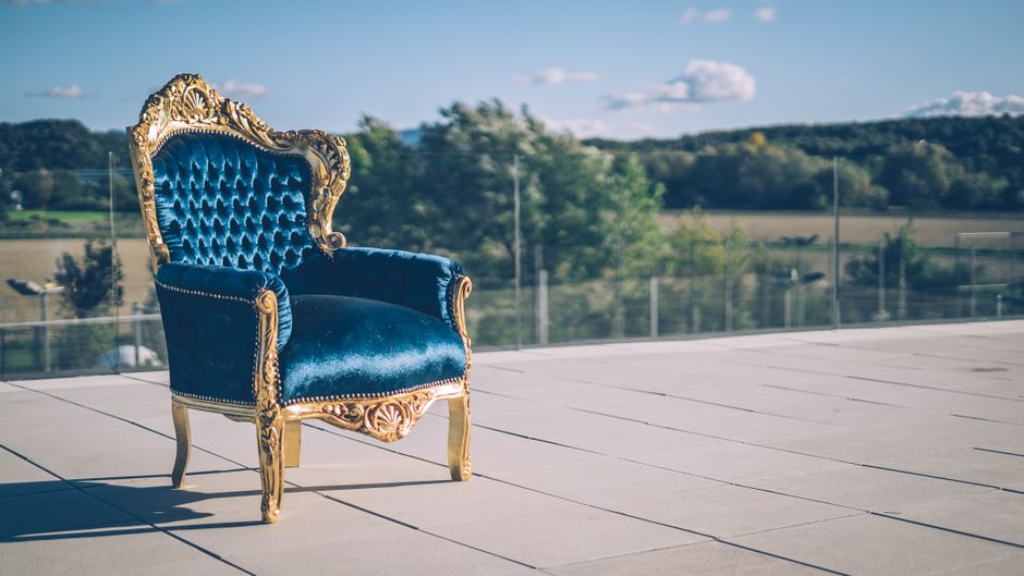 A mustard yellow upholstered armchair with padded armrests and button-tufted backrest sits on small caster wheels, positioned against a plain white wall in a room with a concrete floor. The surface of the chair appears clean and well-maintained, with no visible dust or stains. Natural light illuminates the scene, reflecting subtly on the fabric, indicating a tidy and fresh environment. This image showcases the importance of surface cleaning and maintenance in domestic spaces, aligning with Mile End Park upholstery cleaning tips for Bow residents, SE3, provided by Cleaners E3, specialists in professional cleaning services.