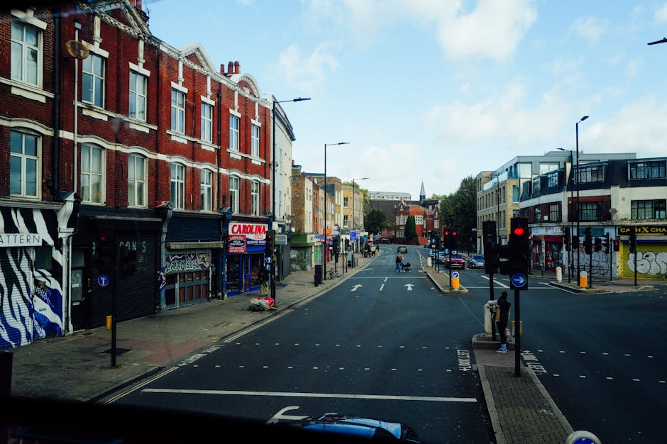 A view of Roman Road Market at the end of the day, featuring a pedestrian crossing with traffic lights, surrounded by a mix of red brick and modern white buildings. The street is lined with various small shops and cafes, some with colorful signage. The pavement appears clean and well-maintained, with street lamps and bollards visible along the sidewalk. The sky above is partly cloudy with patches of blue, and there are a few pedestrians waiting at the crossing. This scene exemplifies an urban commercial area in Bow, SE3, emphasizing the importance of outdoor surface cleaning and maintenance for a tidy, hygienic environment. For professional cleaning services, visit Cleaners E3 for expert support in surface cleaning and sanitisation.