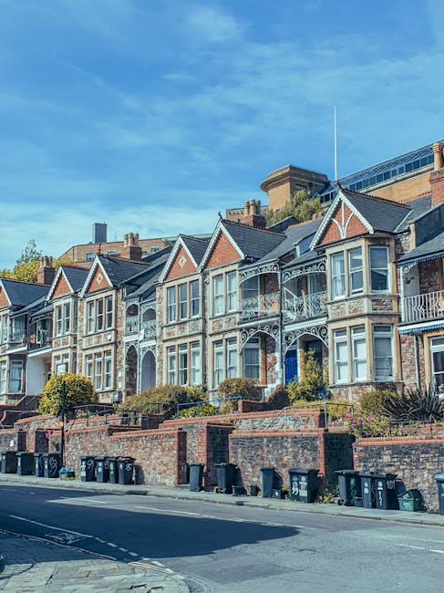 Image of a row of Victorian-style terraced houses with ornate white ironwork balconies and large bay windows, situated along a street with a brick retaining wall and neatly arranged waste bins at the curb. The houses have gabled roofs, some with decorative tiles, and are set against a bright blue sky with scattered clouds. The scene is well-lit, showcasing the clean and maintained facades of the homes. This residential area exemplifies classic British architecture, and Cleaners E3 offers professional cleaning services that include surface cleaning and deep cleaning to maintain such properties’ hygiene and appearance, especially relevant for end of tenancy cleaning in Bow, SE3. The atmospherics highlight the importance of regular maintenance and thorough sanitisation in domestic settings.