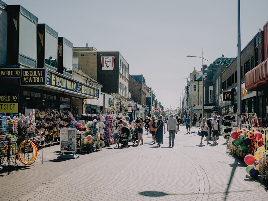 A view of Roman Road Market at the end of the day, featuring a pedestrian crossing with traffic lights, surrounded by a mix of red brick and modern white buildings. The street is lined with various small shops and cafes, some with colorful signage. The pavement appears clean and well-maintained, with street lamps and bollards visible along the sidewalk. The sky above is partly cloudy with patches of blue, and there are a few pedestrians waiting at the crossing. This scene exemplifies an urban commercial area in Bow, SE3, emphasizing the importance of outdoor surface cleaning and maintenance for a tidy, hygienic environment. For professional cleaning services, visit Cleaners E3 for expert support in surface cleaning and sanitisation.